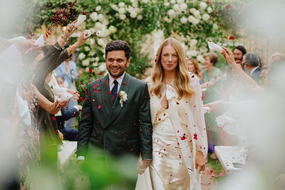Newlyweds walk through garden as guests throw colorful confetti. Bride in white slip dress, groom in dark green suit under archway