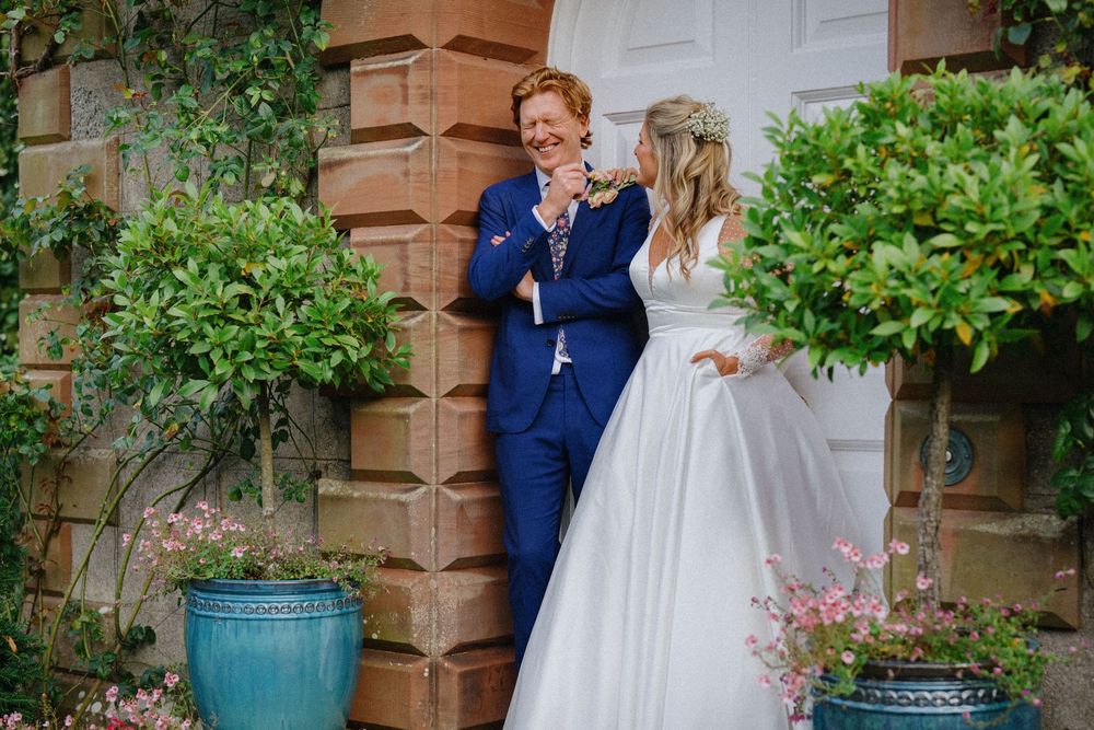 Bride & groom outside brick building with white door and ivy-covered walls. One in navy blue outfit, other in wedding dress