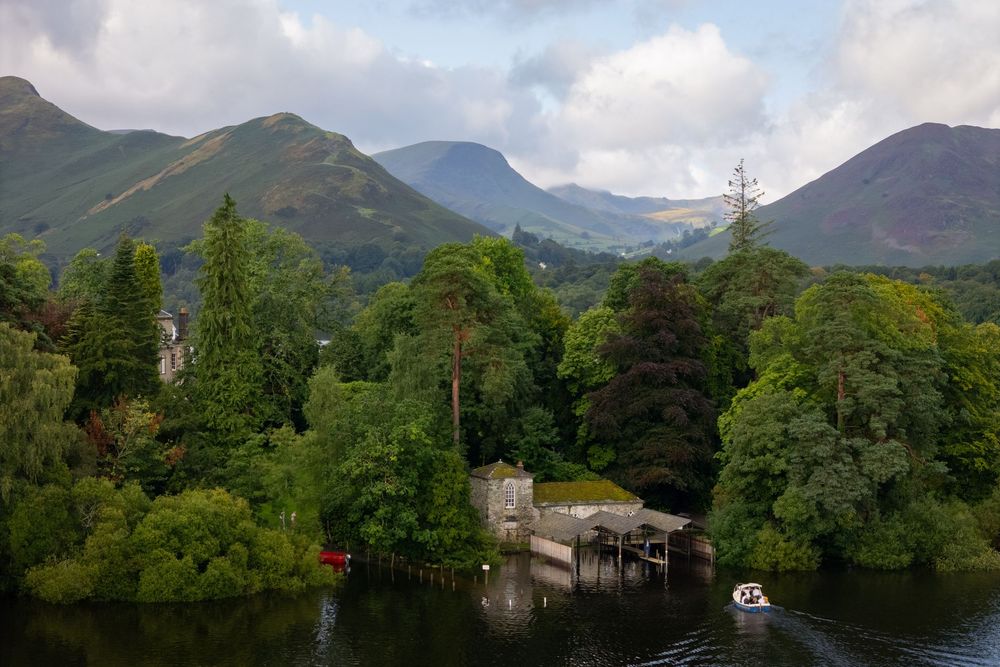 Scenic lake view with rolling green hills & mountains in background. Stone boathouse and jetty visible among dense woodland under cloudy sky