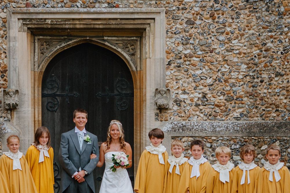 Bride & groom with children's choir in yellow robes standing before medieval church entrance. Ancient stone and flint wall with Gothic door