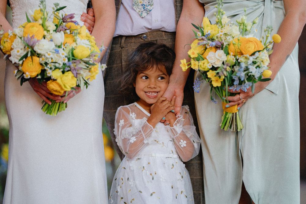 Adorable flower girl in white dress with star pattern poses between bridesmaids in white dresses hold yellow and white bouquets