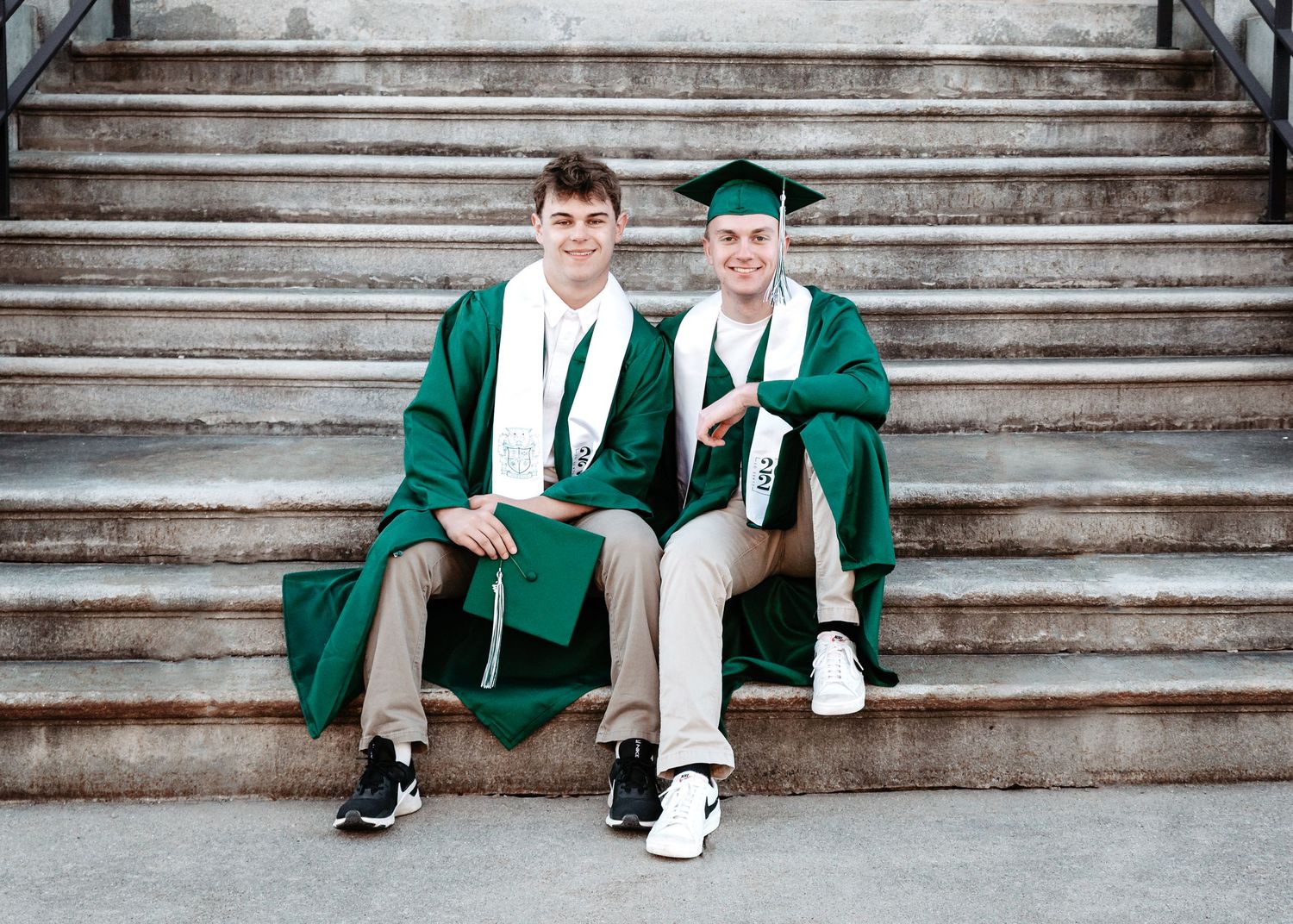 Two graduates in green caps and gowns sit together on stone steps during commencement ceremonies.