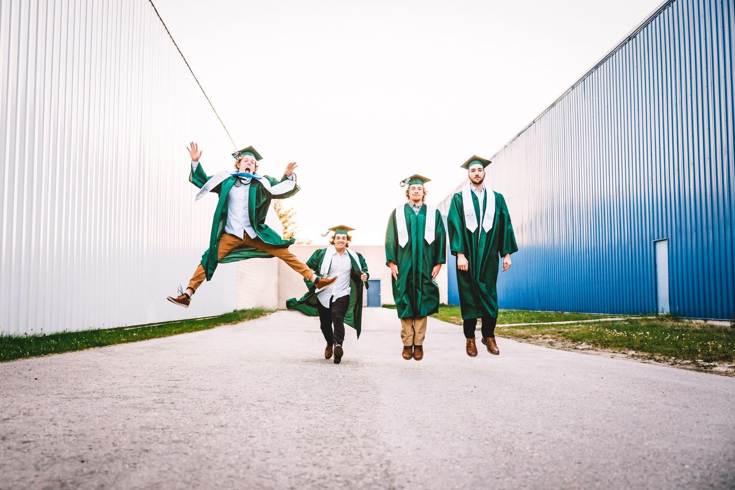 A group of graduates in green caps and gowns walk together on a path while one jumps joyfully for a photo.