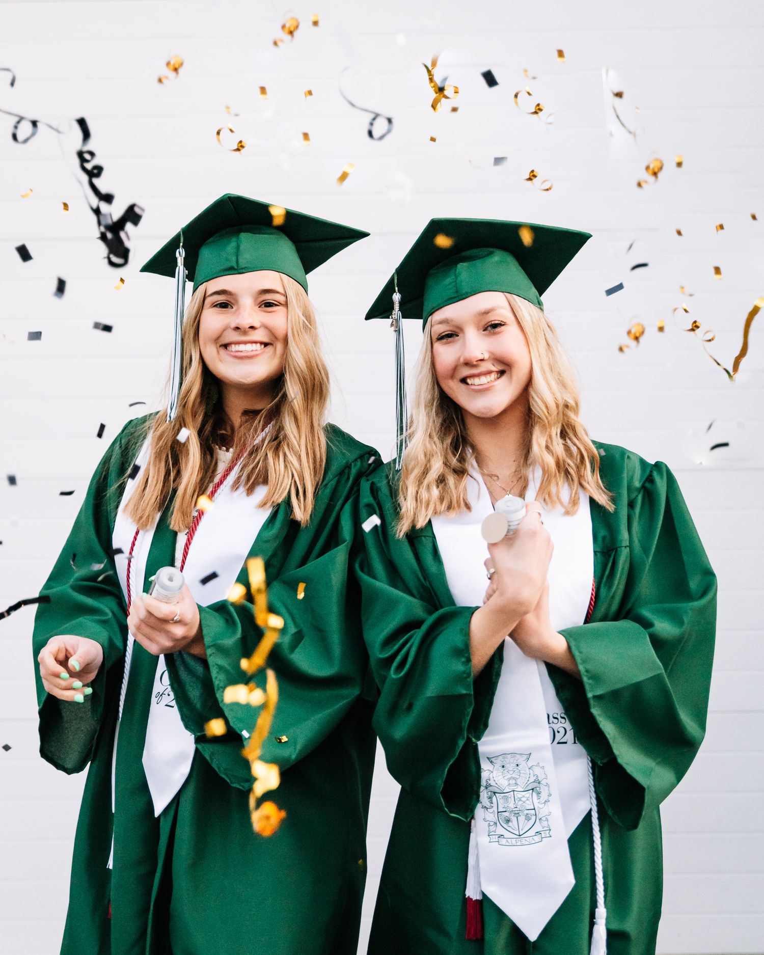 Two graduates in green caps and gowns celebrate with confetti falling around them against white background.