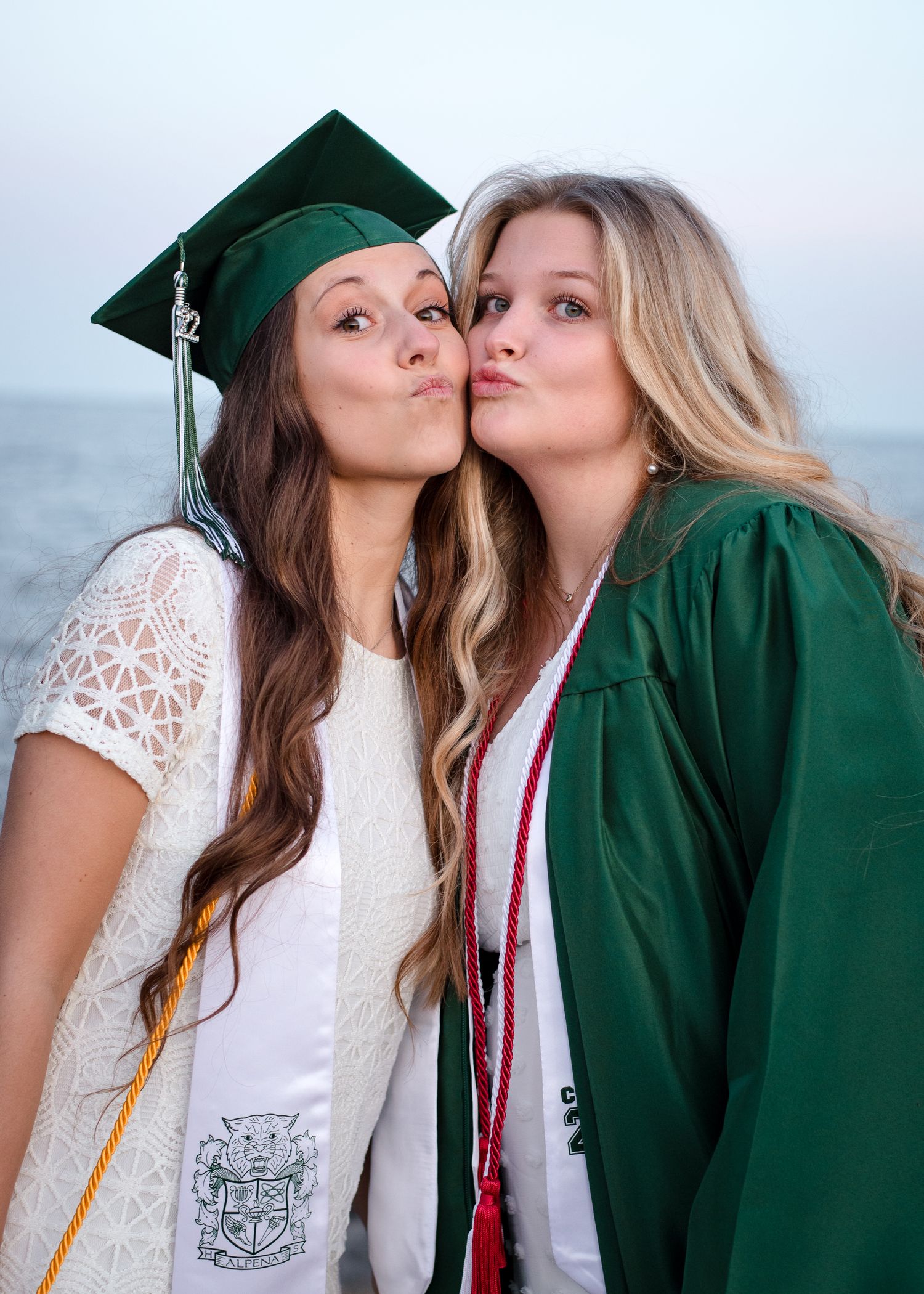 Two people in green graduation caps and gowns posing for a playful photo outdoors on graduation day.