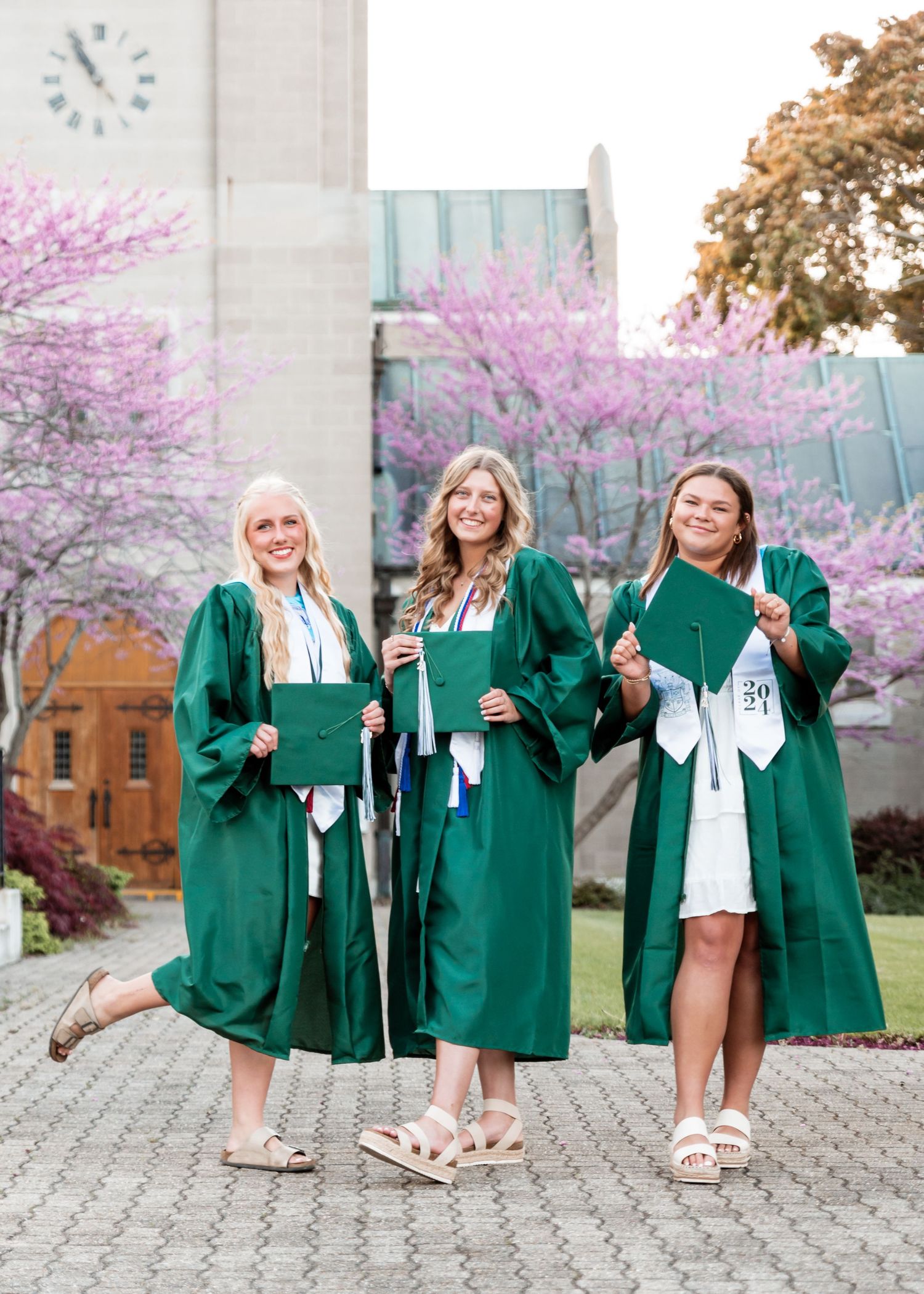 Three graduates wearing green commencement robes and holding diplomas pose together under blooming cherry trees.