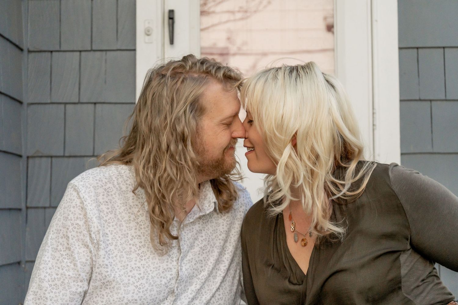 Romantic couple sharing an intimate moment, touching foreheads and almost kissing outside a gray shingle home.