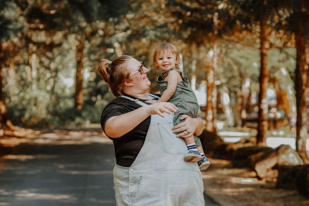 A mother in overalls lifts her smiling toddler in a sunny tree-lined park path.