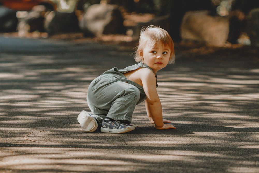 An adorable toddler in green overalls crawling on a sunlit pavement outdoors.