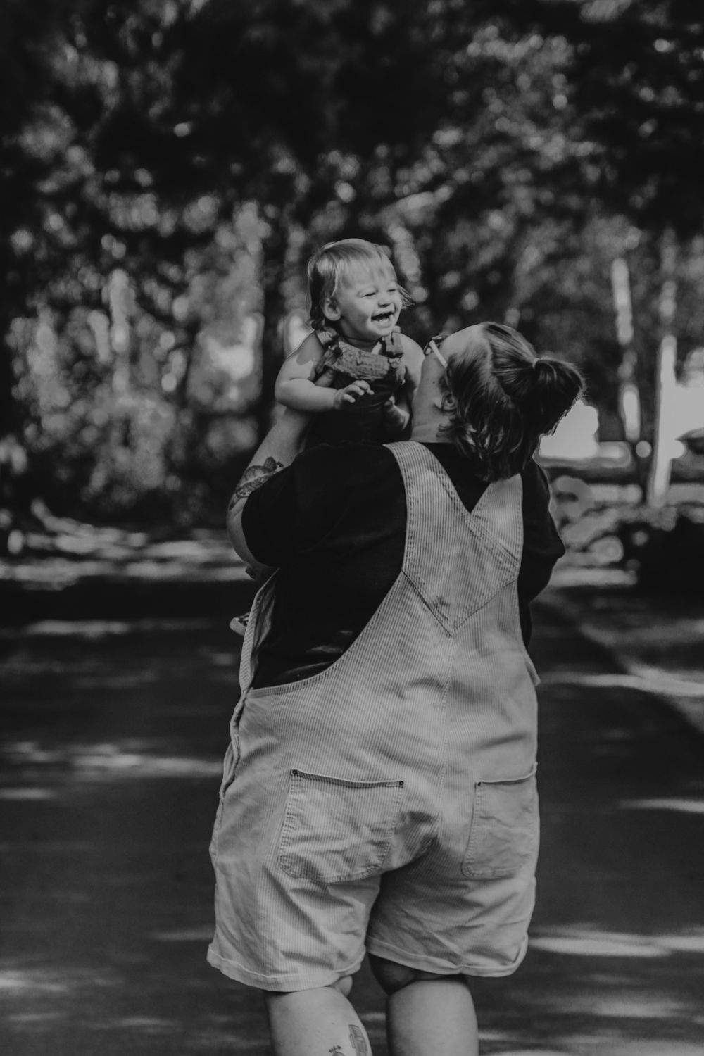 Black and white photo of a mother lifting her laughing toddler in a tree-lined park.