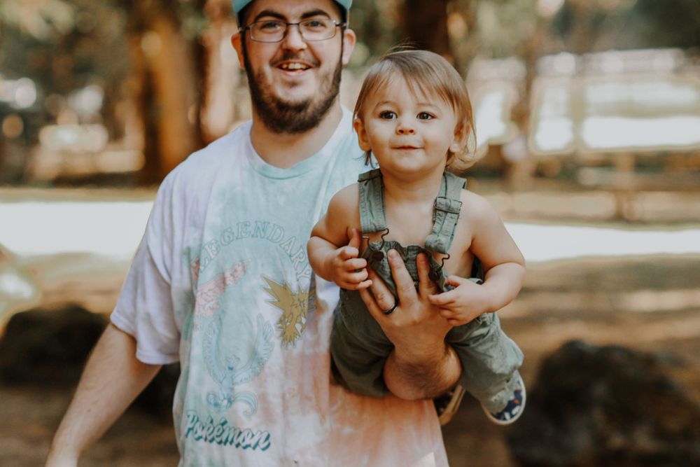 A smiling father in a tie-dye shirt holds his happy toddler in a sunlit park.