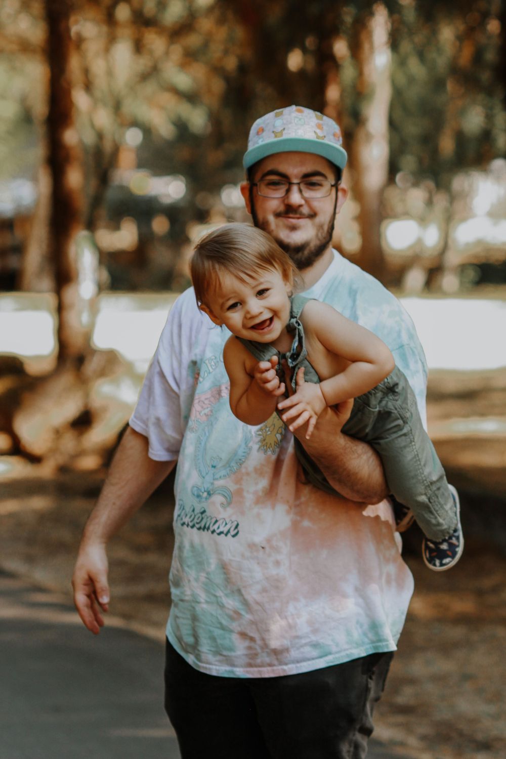A dad in a tie-dye shirt and green cap carries his laughing toddler in a park.