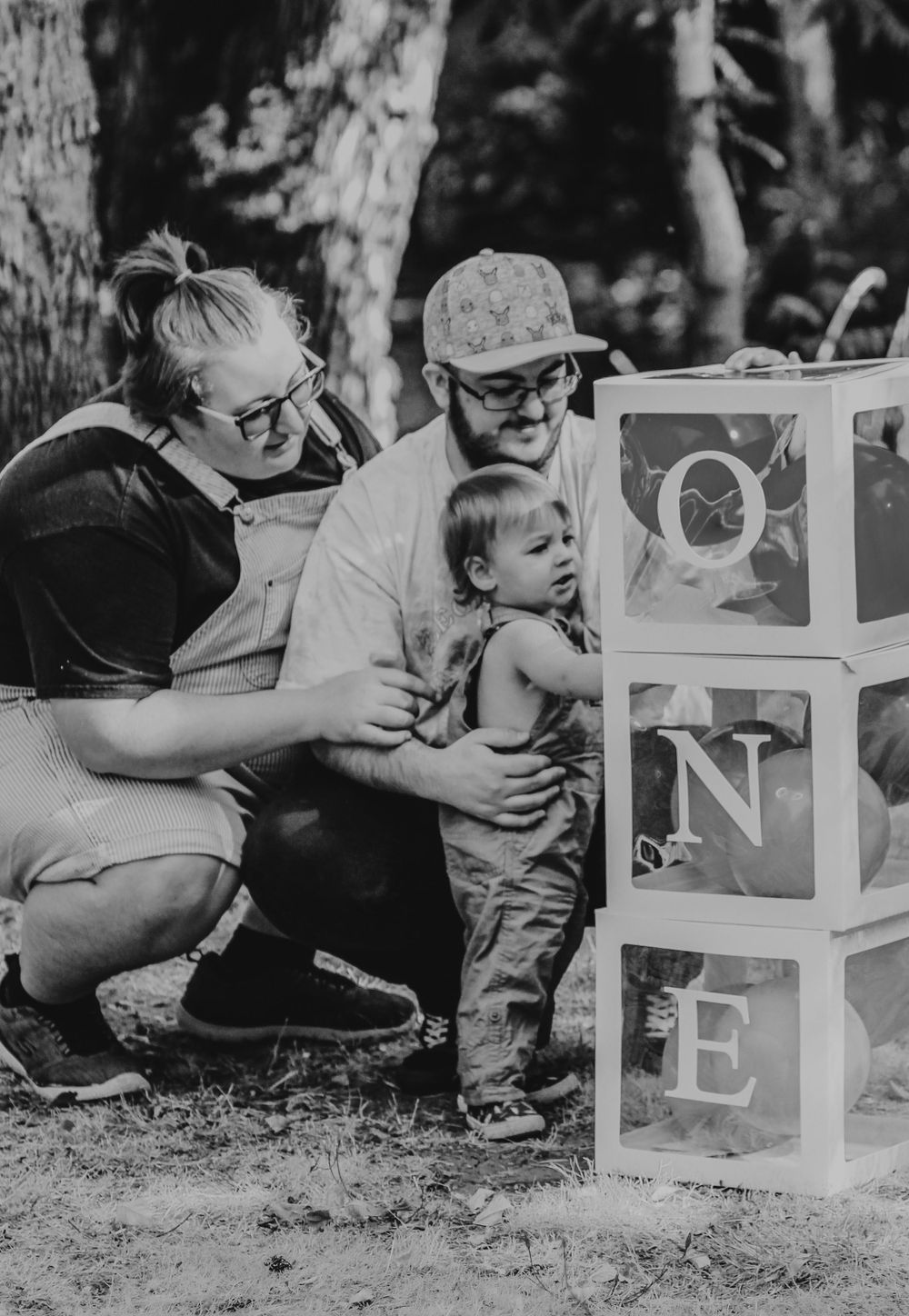 Black and white photo of parents crouching with their toddler near letter blocks spelling ONE.