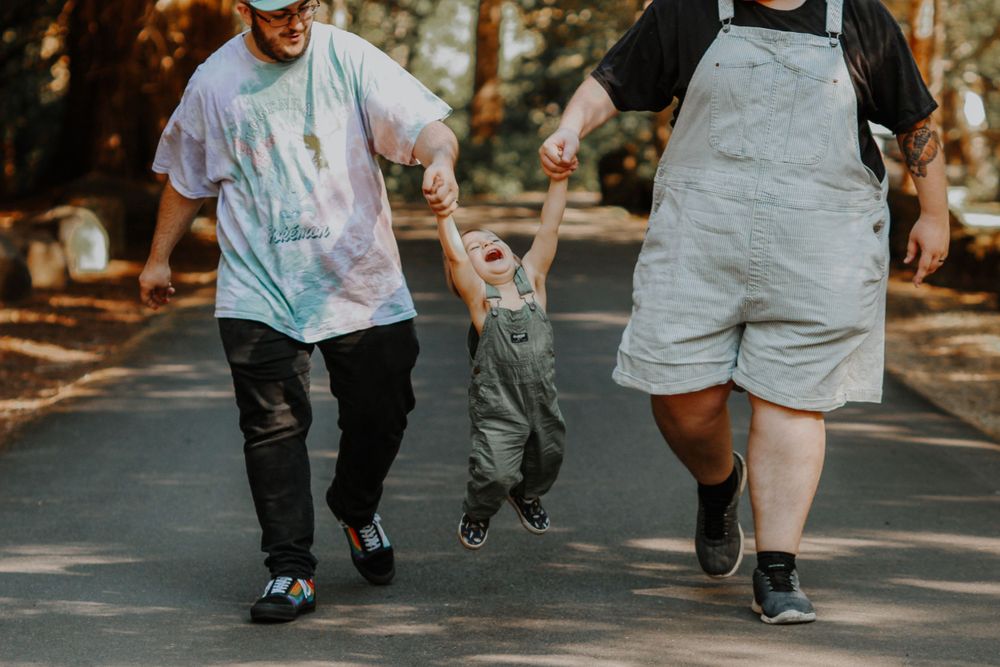 Two parents swing their toddler by the hands while walking down a sunny tree-lined path.