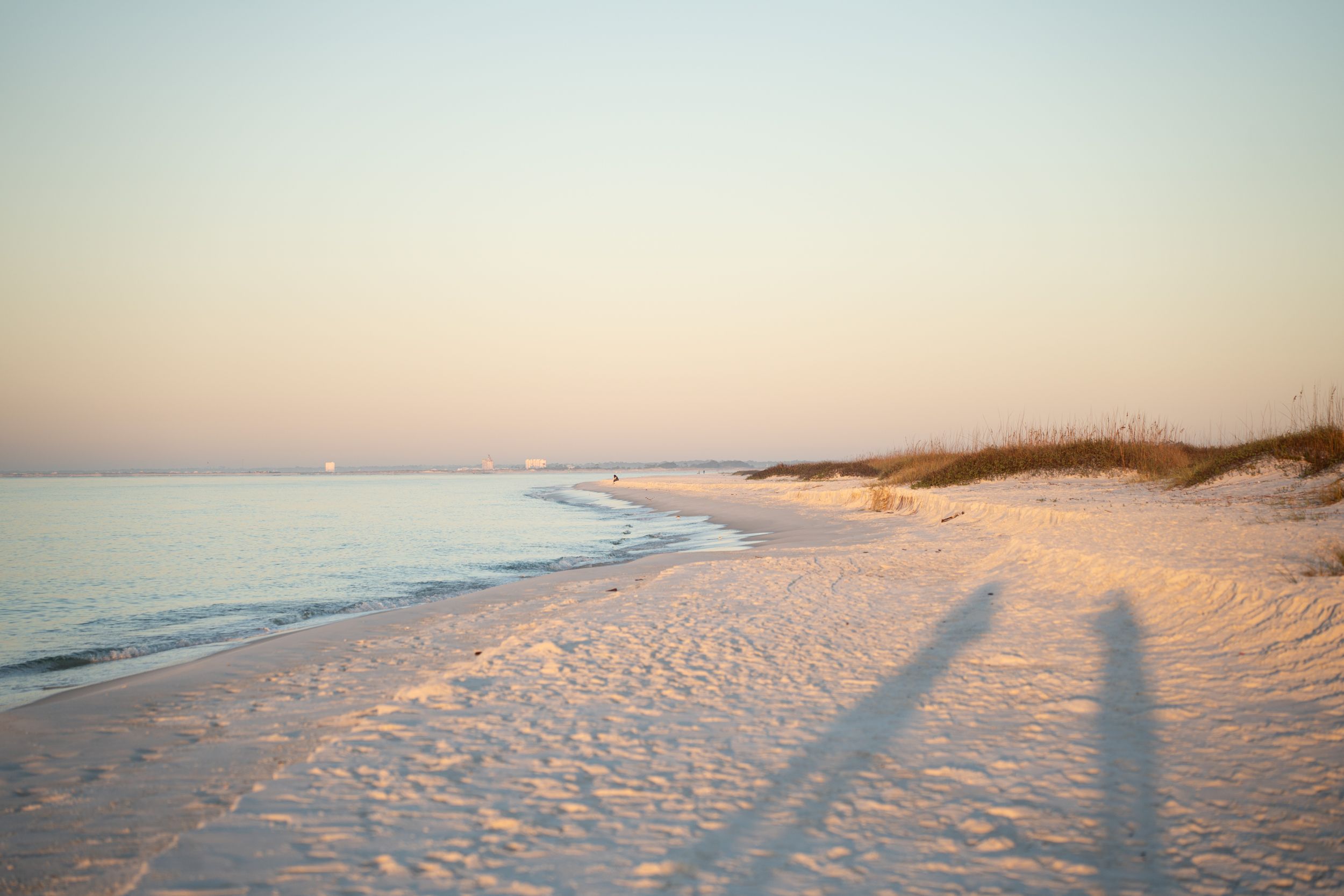 Long shadows stretch across pristine white sand beach at sunset with gentle waves rolling onto shore at dusk.