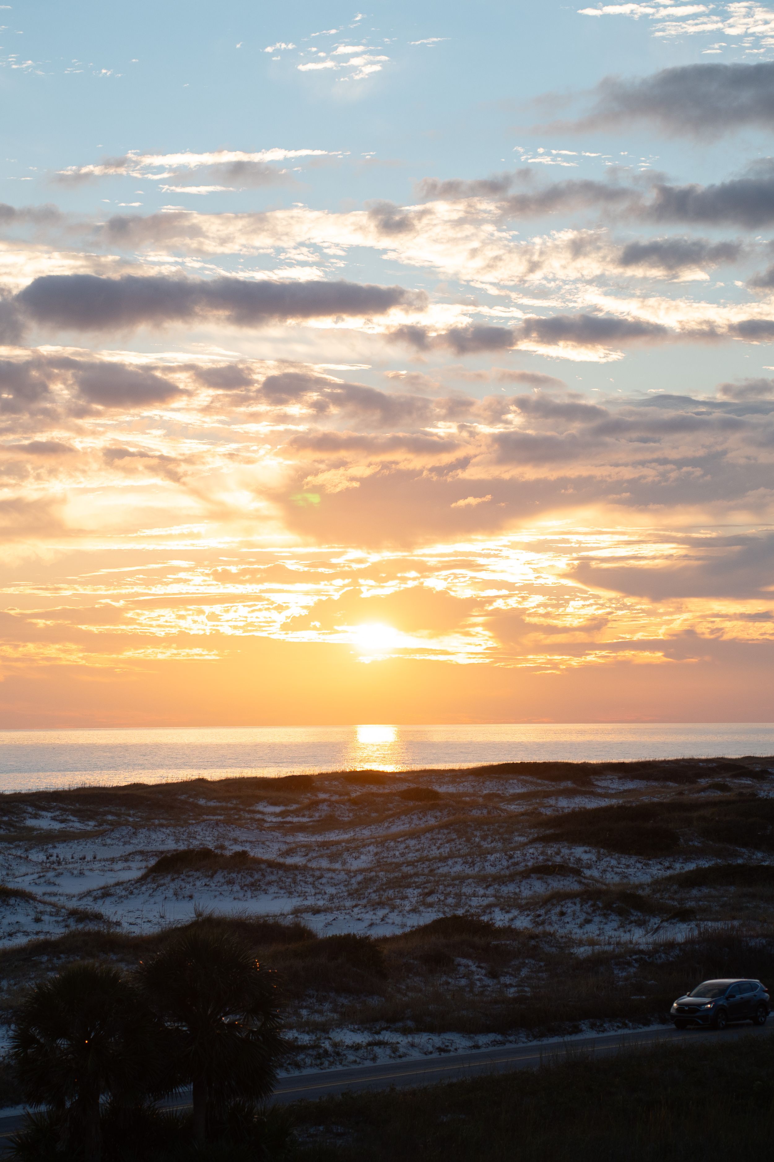 Dramatic orange sunset over coastal dunes with clouds reflecting golden light across the horizon.