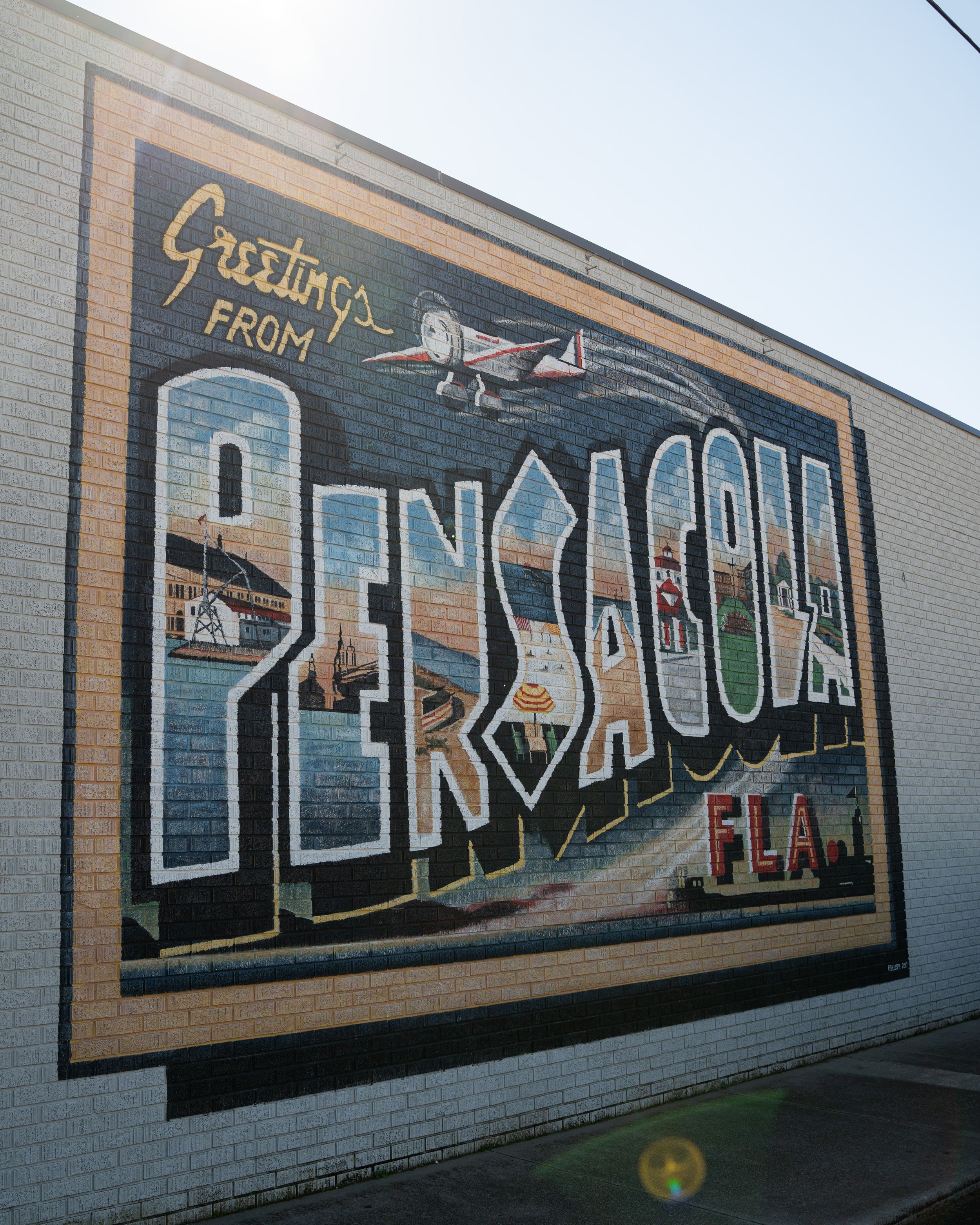 A vintage-style mural on a brick wall displays 'Greetings from Pensacola' in large decorative lettering.