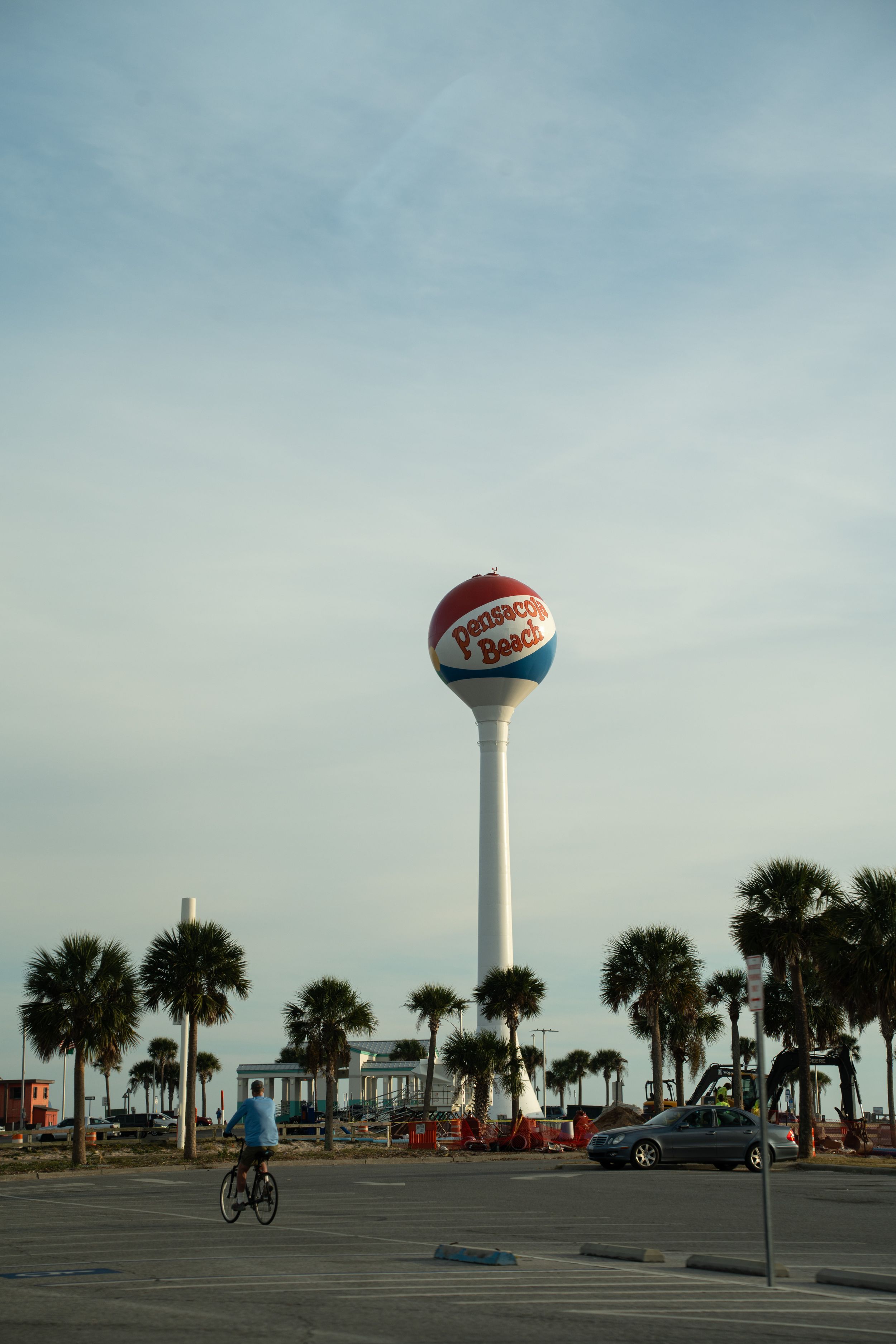 A Pepsi-Cola water tower rises above palm trees in a beachfront parking lot with a cyclist riding by.