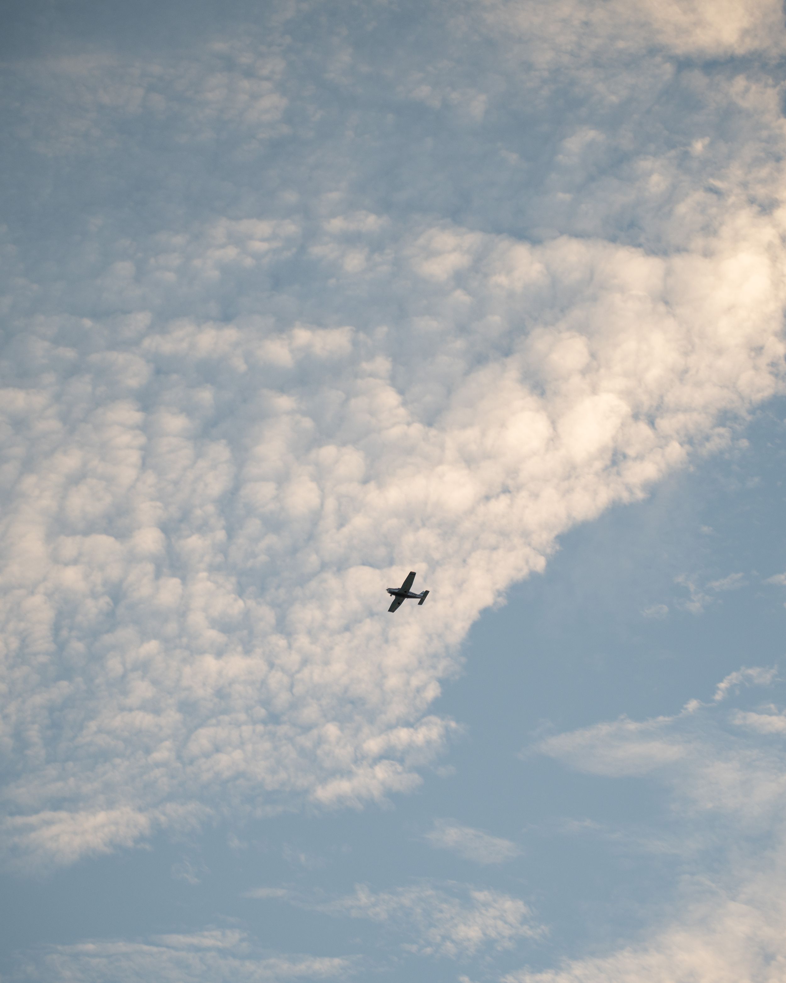 Small airplane flies through cloudy blue sky with scattered altocumulus clouds stretching across frame.