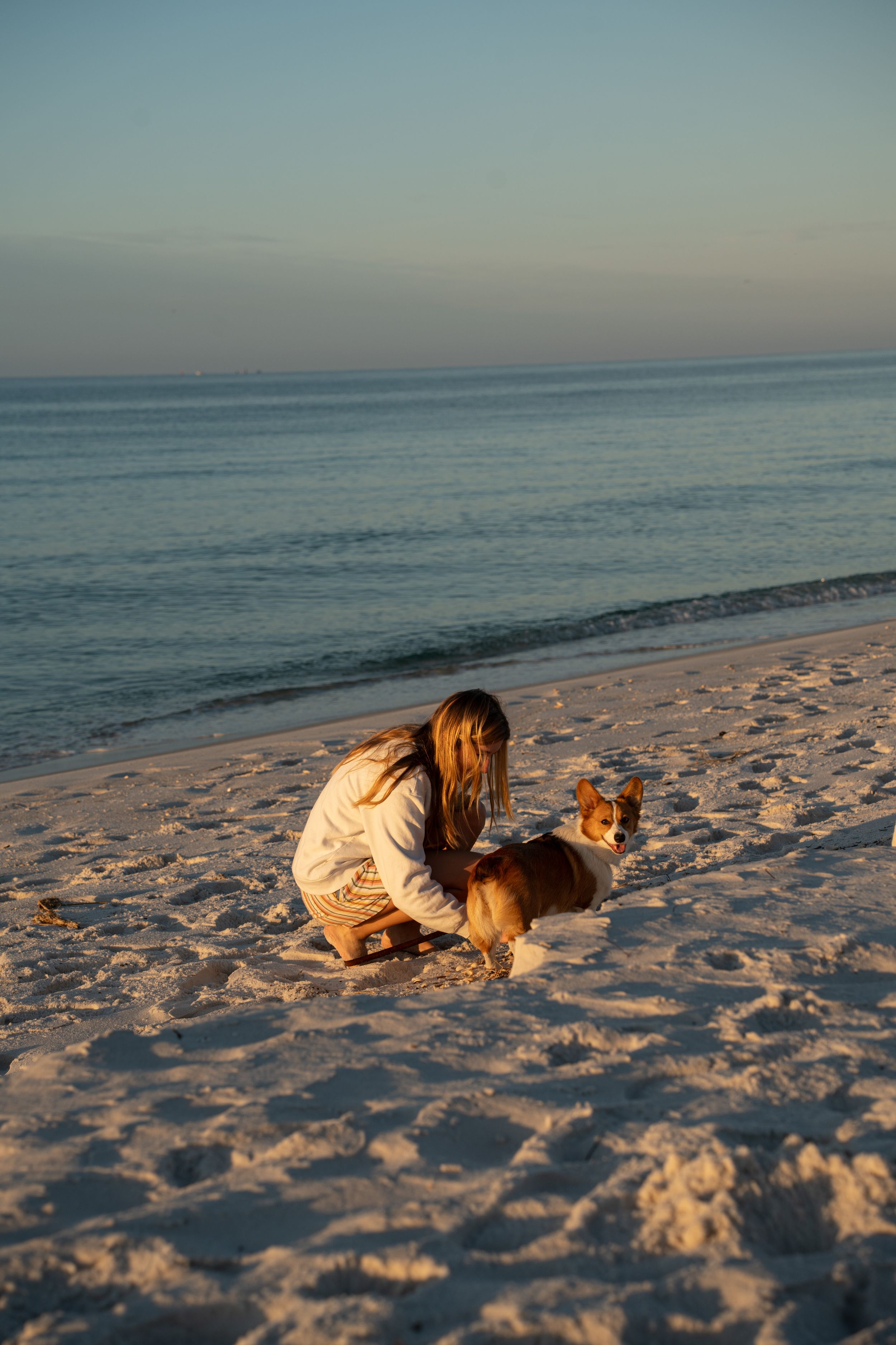 Someone in a tan outfit kneels on a sandy beach with a golden retriever dog during sunset by the ocean.