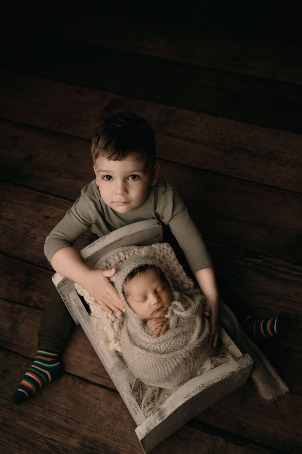 A child in striped socks relaxes on wooden floor beside newborn baby sleeping in cozy knit wrap.