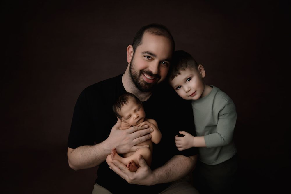 Family portrait in muted tones against dark background showing an adult holding a baby with a child beside them.