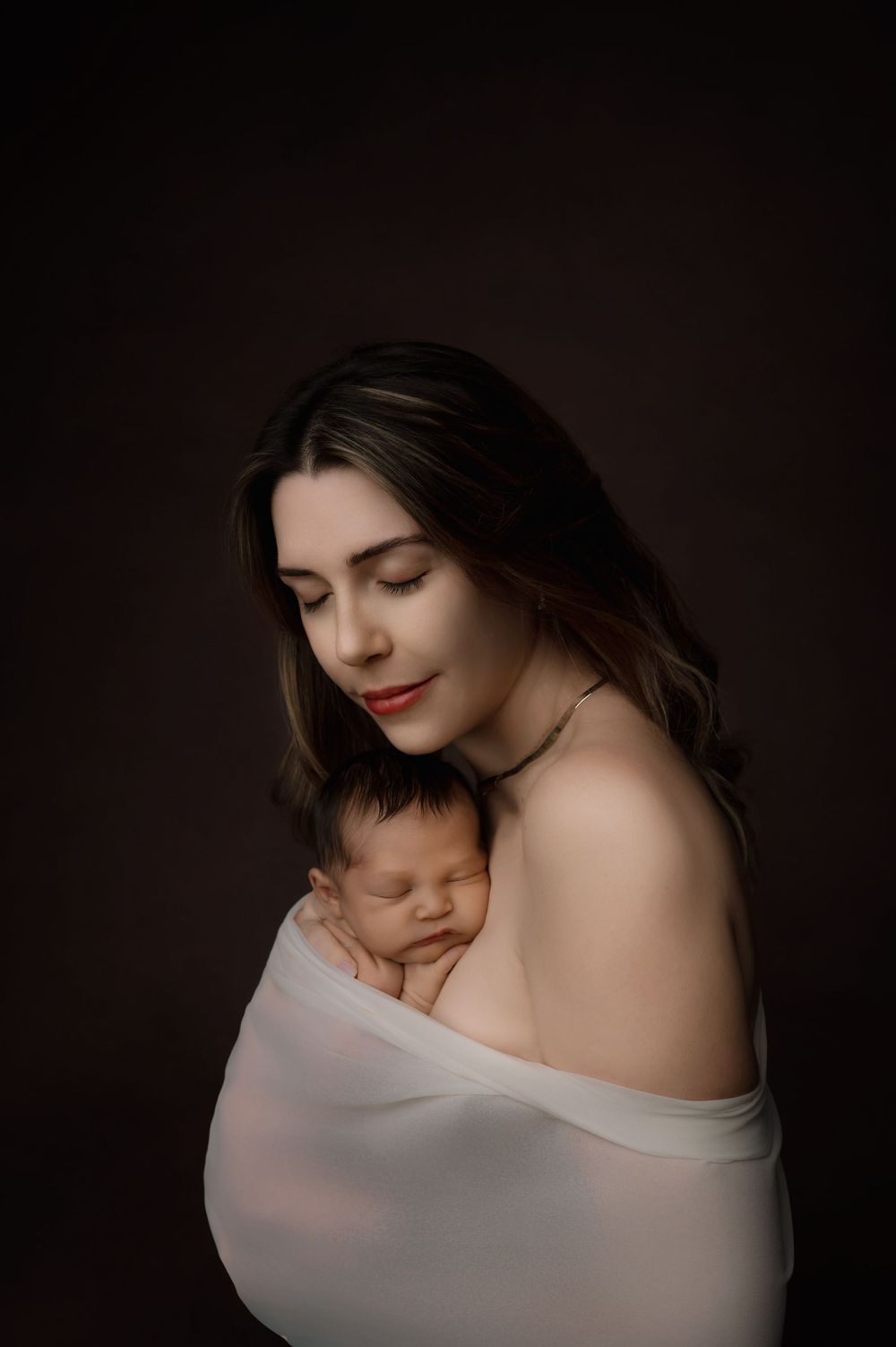 A tender motherhood portrait in dark tones shows a woman holding a newborn baby wrapped in soft white fabric.