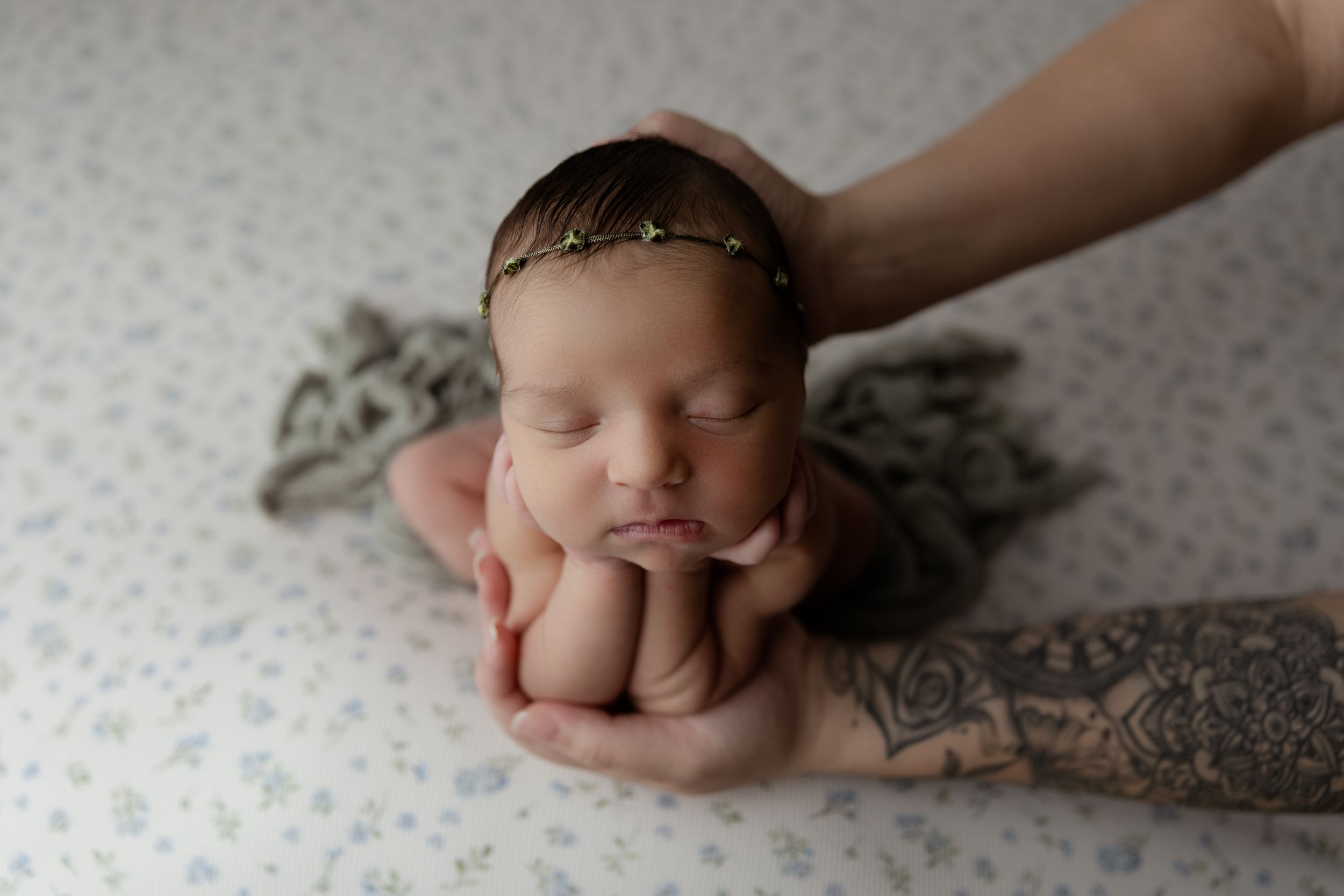Sleeping newborn supported by a tattooed arm in froggy pose on a floral backdrop.
