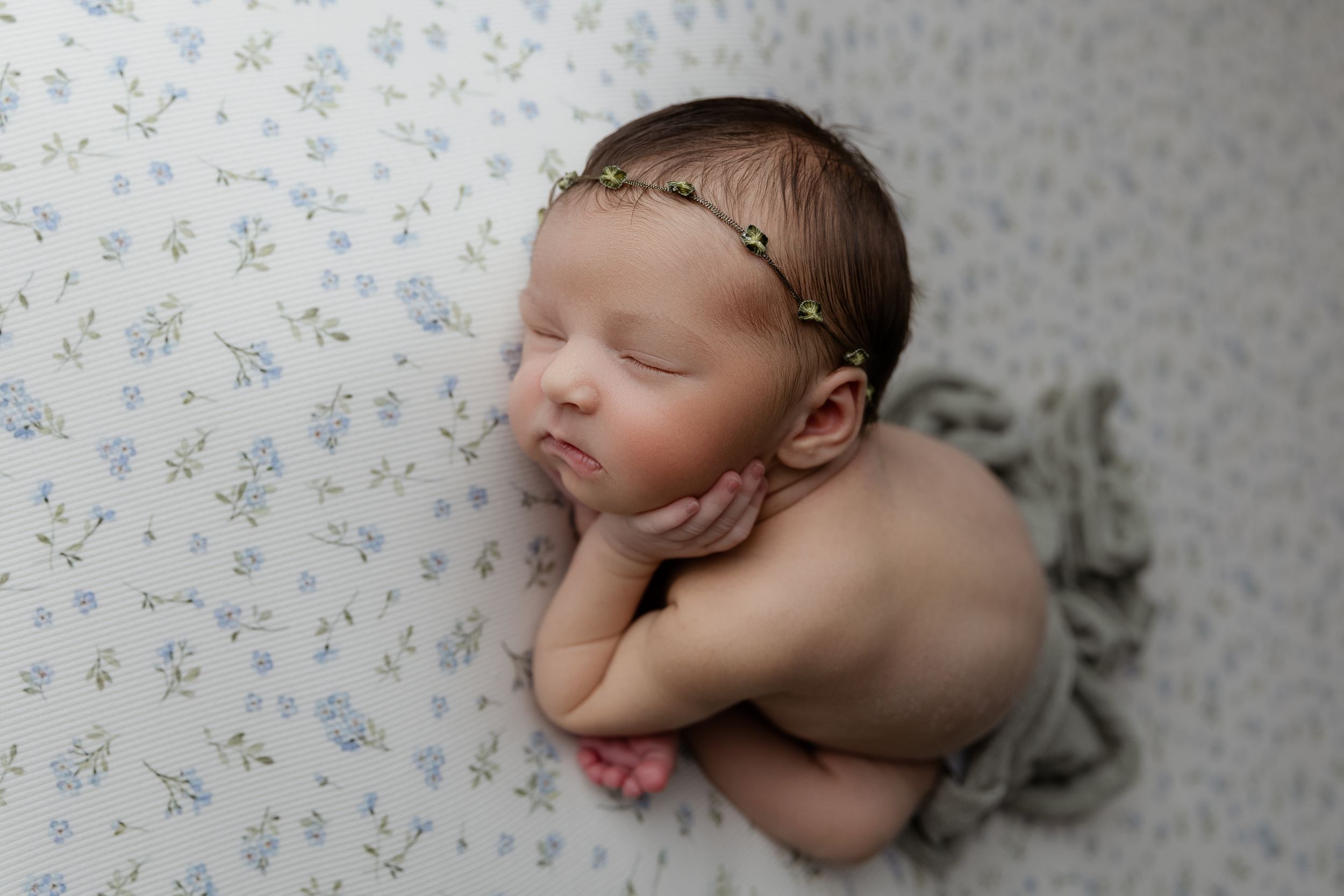 Peaceful sleeping newborn baby laying on floral print fabric wearing delicate leaf crown headband.