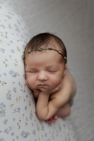 Close-up portrait of sleeping newborn baby wearing green leaf crown headband on floral patterned blanket.