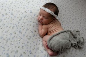 Newborn baby sleeps on floral patterned blanket wearing delicate white headband and gray wrap.