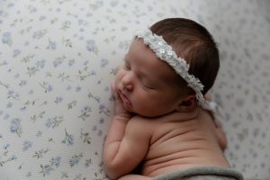Sleeping newborn wearing delicate white lace headband rests on floral pattern fabric.