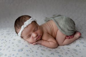 Newborn baby wearing delicate lace headband sleeps peacefully on floral fabric while wearing gray diaper cover.