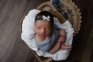 Sleeping newborn wrapped in soft blue cloth lies peacefully in brown woven basket on white blanket.