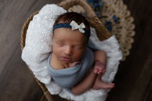 Newborn wrapped in blue swaddle resting in basket with white blanket and bow headband.