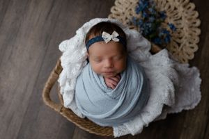 A sleeping newborn wrapped in soft blue fabric lies in a woven basket on white lace with blue flowers in background.