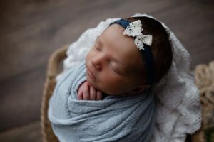 Peaceful newborn swaddled in gray wrap sleeping in wooden basket with delicate bow headband.