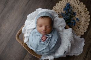 Newborn sleeping peacefully in a basket lined with white lace while swaddled in light blue fabric.