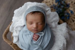 A sleeping baby wrapped in a soft blue swaddle blanket and white bonnet rests in a cozy basket.