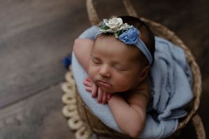 A sleeping baby rests in a woven basket wearing a floral headband and draped in blue fabric.