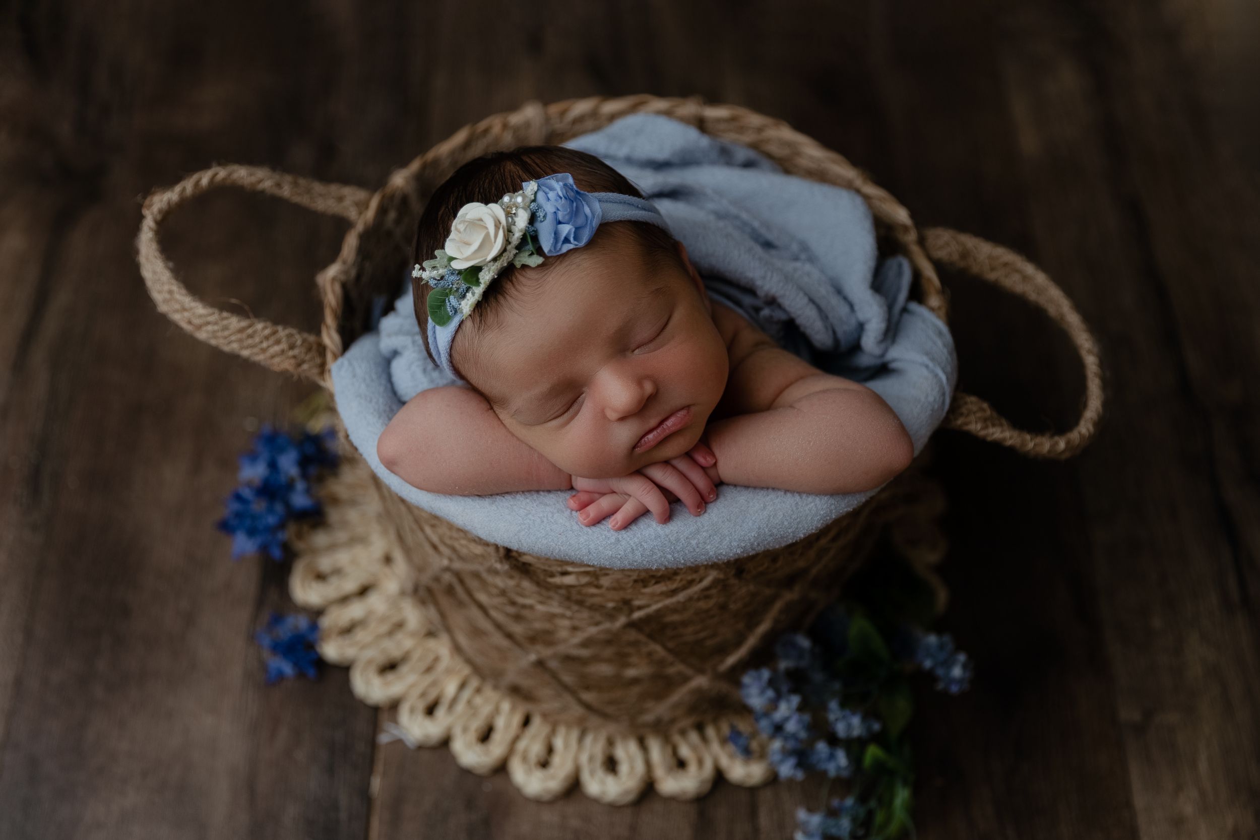 Sleeping newborn wearing light blue wrap rests peacefully in rustic basket surrounded by blue flowers on wooden floor.