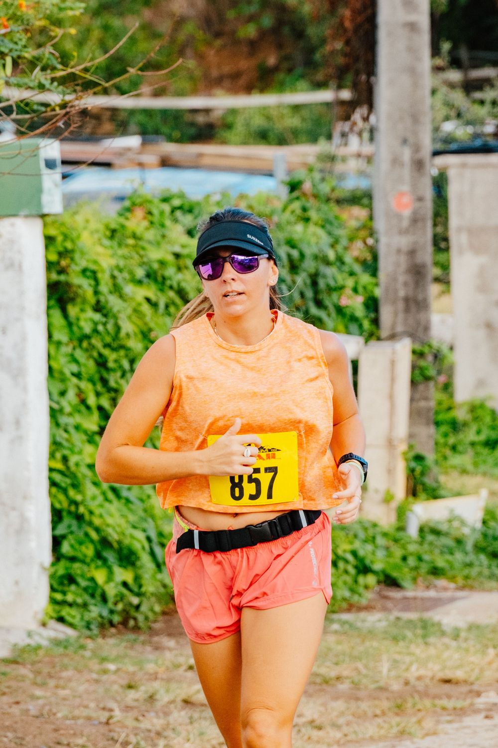 A runner in an orange tank top and coral shorts participates in a race wearing number 857 on a dirt path.