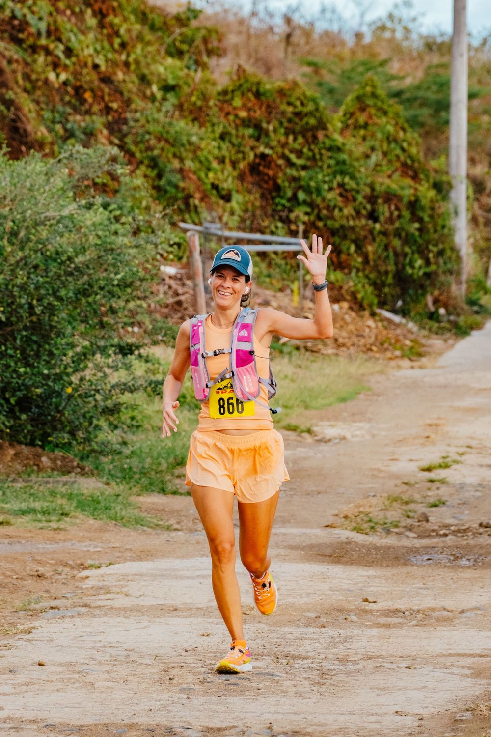 Runner in bright athletic wear and race number waves while jogging along a dirt trail surrounded by greenery.