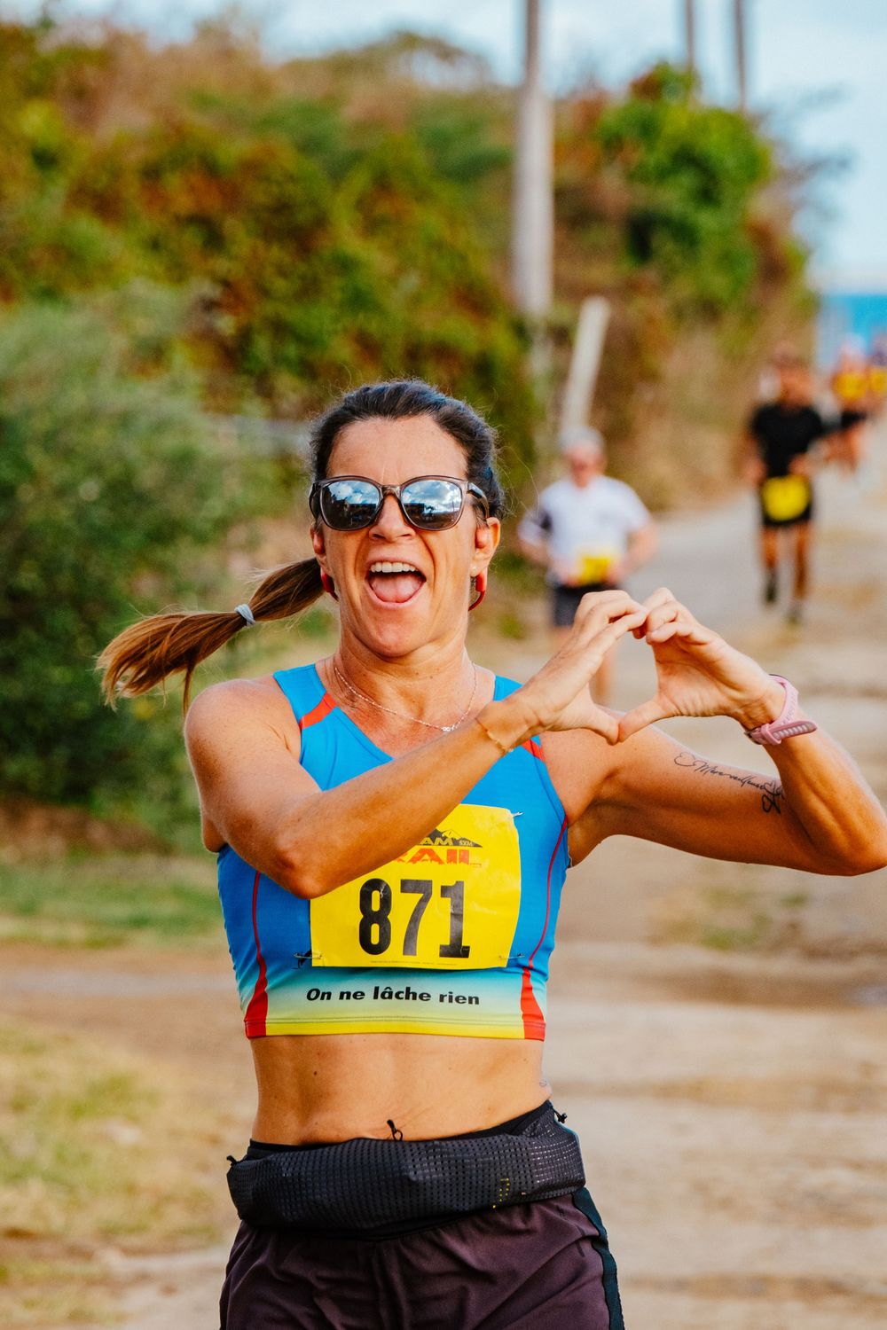 Runner with bib number 871 in a blue tank top shows enthusiasm during outdoor race on sunny trail path.