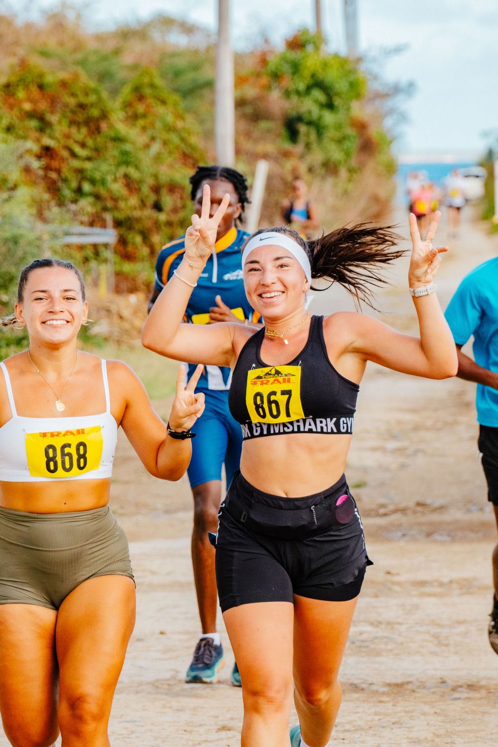 Happy runners in athletic wear smiling and making peace signs during outdoor race event on dirt path near beach.