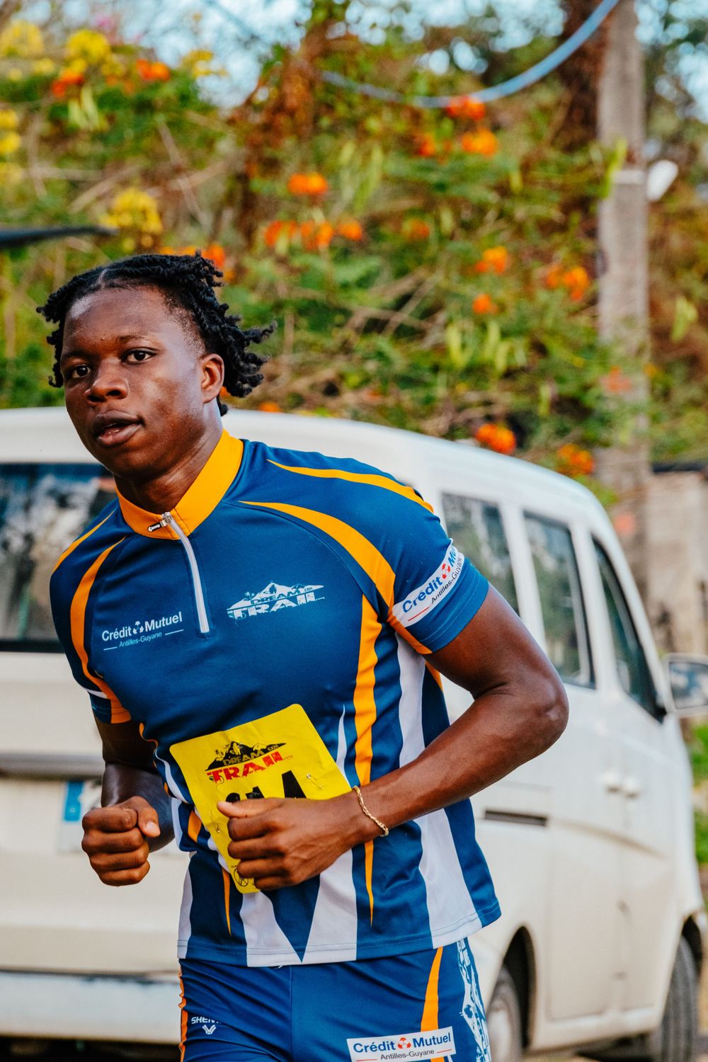 Athletic runner in blue and yellow racing uniform moves past parked car with orange flowers in background.