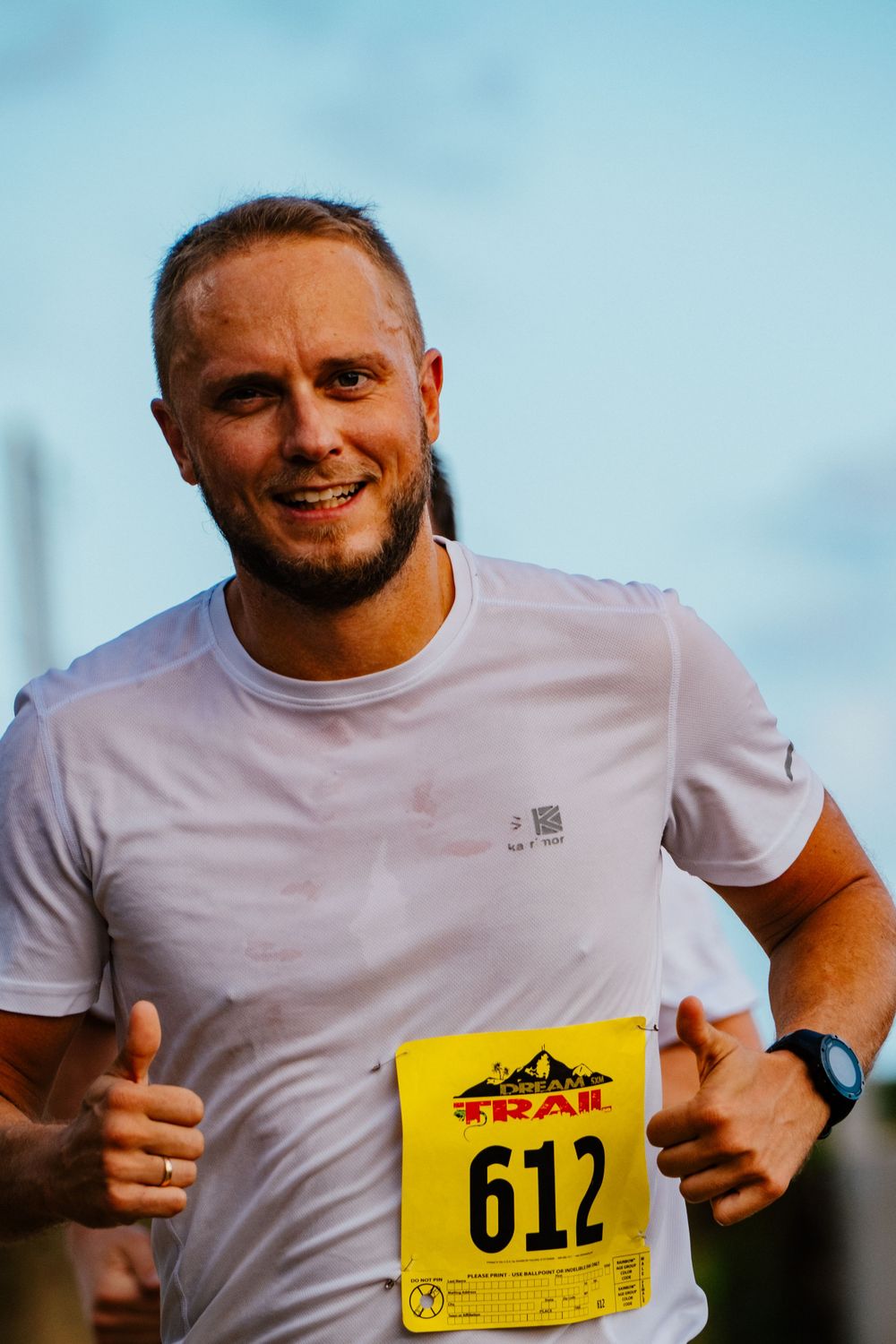 Runner wearing a white t-shirt and race number participates in an outdoor competition against blue sky background.