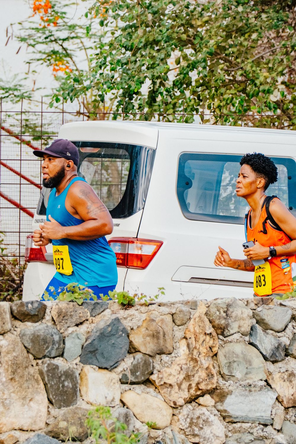 Two runners in blue athletic attire race along a stone wall with a white vehicle parked behind them.