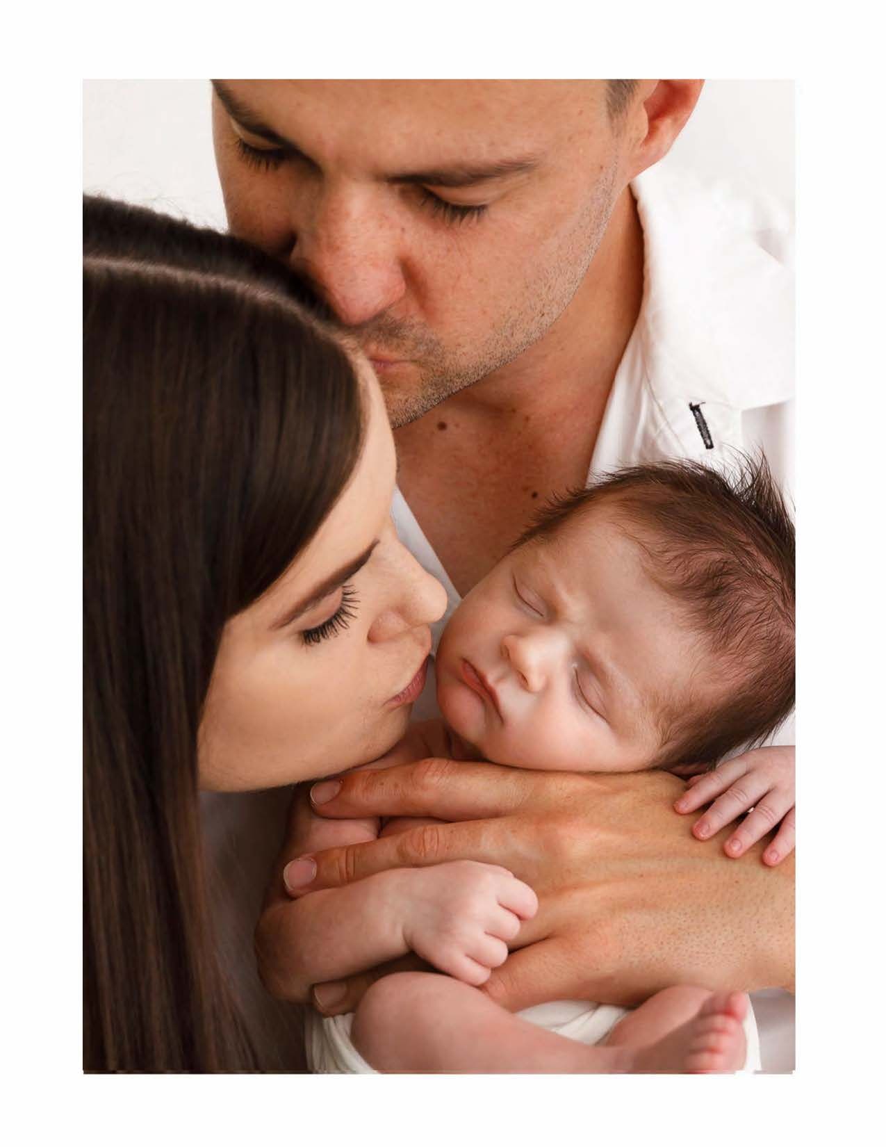 A tender family portrait showing parents sharing an intimate moment with their sleeping newborn baby in warm lighting.