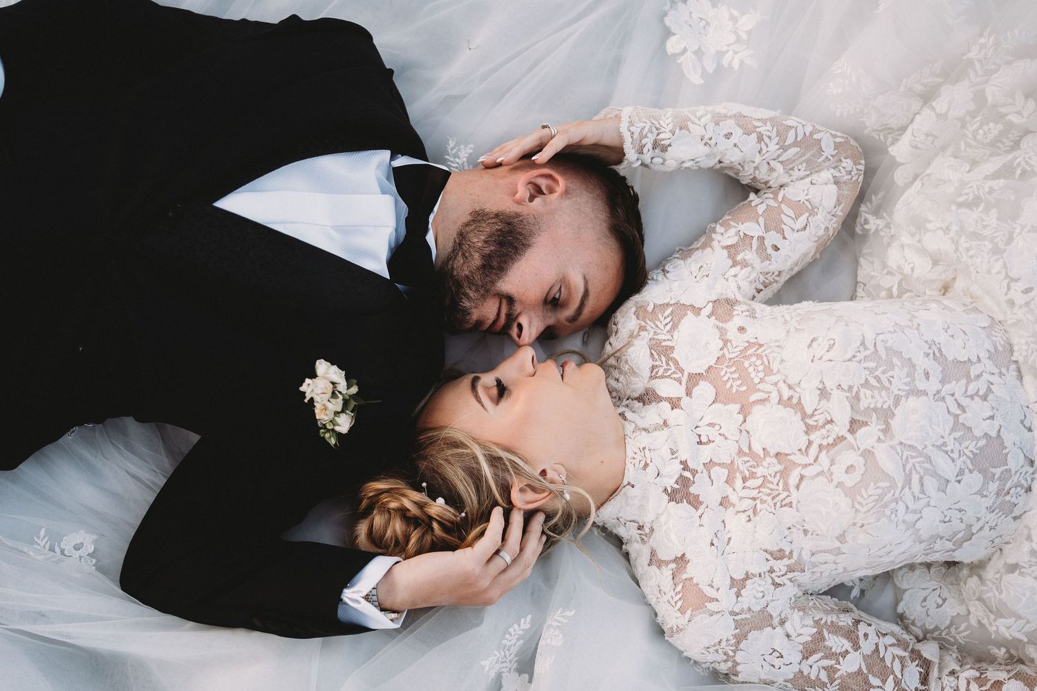 A bride in white lace dress and groom in black suit lying together on white background.